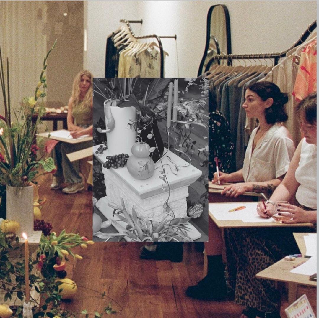 A session in a clothing showroom: women seated at small wooden tables with sheets of white paper and coloured pencils, a glass of rosé, a strip of colour swatches. Racks of garments on gold rails behind them, a tall arched mirror, a lit taper candle and a large arrangement of flowers and fruit in the foreground. A black-and-white photograph of the staged still life — a white jug, tomatoes, grapes and a candle on a carved stone plinth — is laid over the centre.