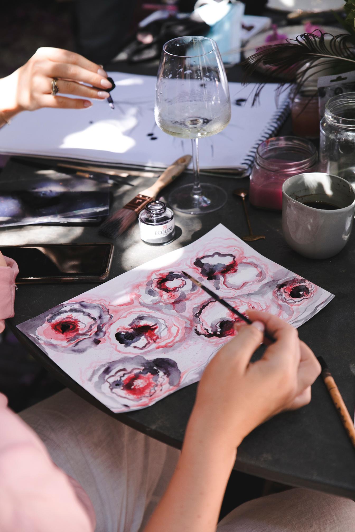 Close view of two pairs of hands working at a table. In front, a brush is laid across a sheet of paper covered with six loose blooms in crimson and dark ink. Beside it, an open bottle of coloured drawing ink, a wide flat brush, jars of clear and pink-tinted water, a coffee cup and a half-drunk glass of white wine.