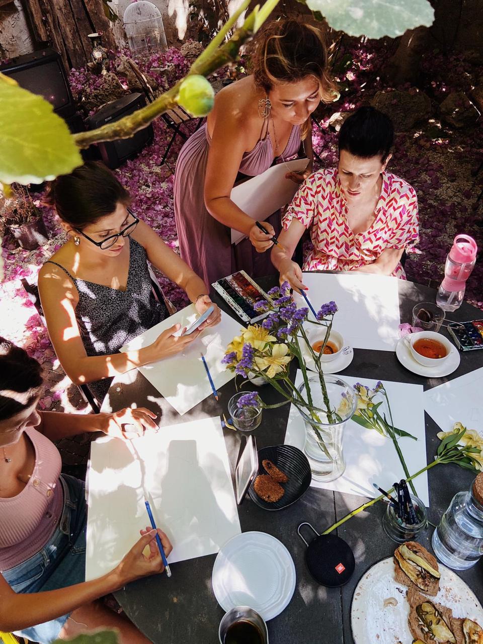 Four women at a round stone table in a garden, seen from above. Large sheets of white paper in front of each of them, still nearly empty. An open box of watercolour pans, blue pencils, brushes in a glass, cups of tea, biscuits and open sandwiches, a vase of purple and yellow flowers, and a small speaker on the table. Dappled sunlight and a bank of pink bougainvillea behind.