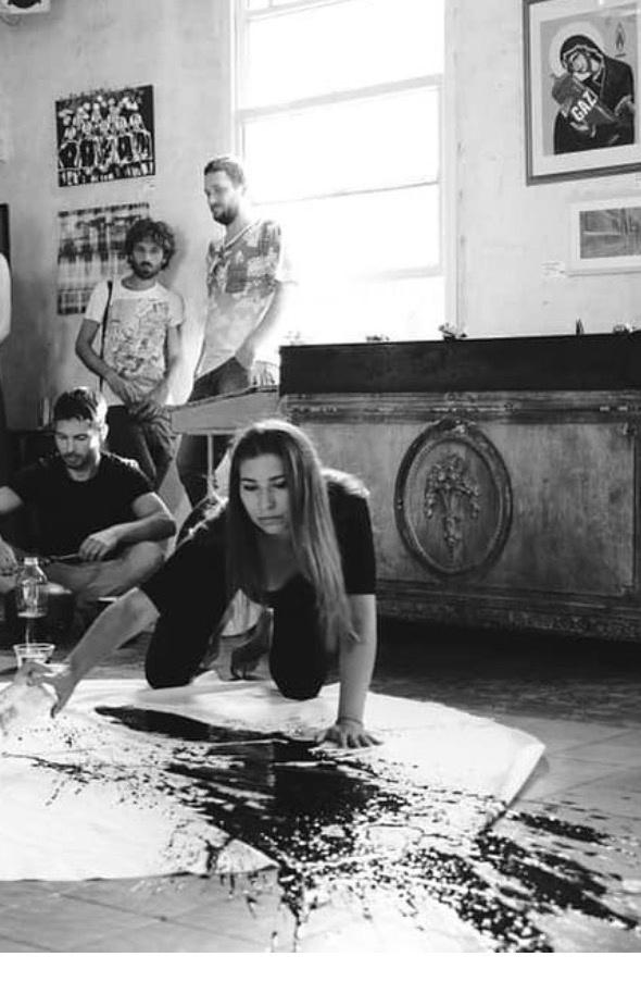 Black and white photograph. Karina Cohen kneels on a large sheet of paper spread across a tiled floor, one hand planted on it, the other arm extended, mid-gesture. Black ink is thrown and pooled across the paper. Three men watch from the background of the room, beside a tall window and a heavy carved sideboard.