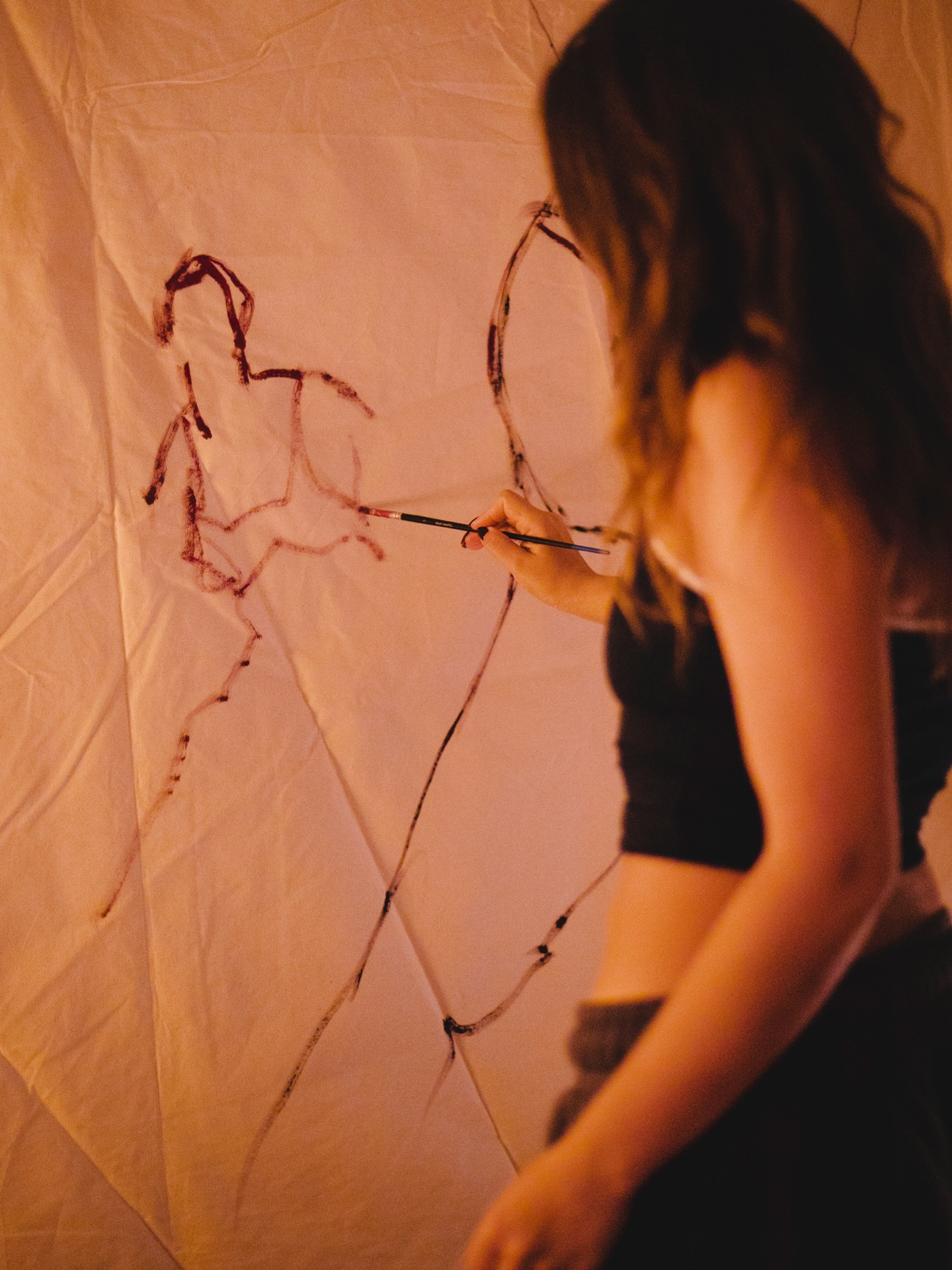 Karina Cohen painting continuous dark red figures onto a large white cloth hung on a wall, brush in hand, seen from behind in warm low light.