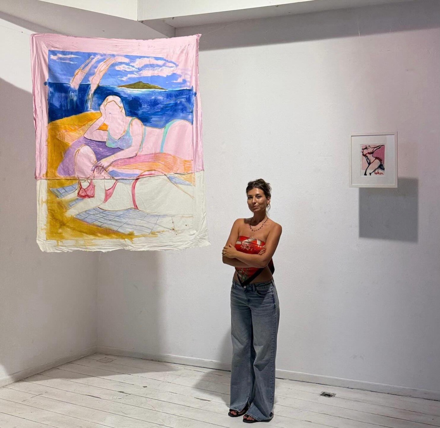 A painting on unstretched cloth with a soft pink border, hung suspended in a white gallery with painted floorboards. A small framed figure study hangs on the wall beside it, and Karina Cohen stands in the room.